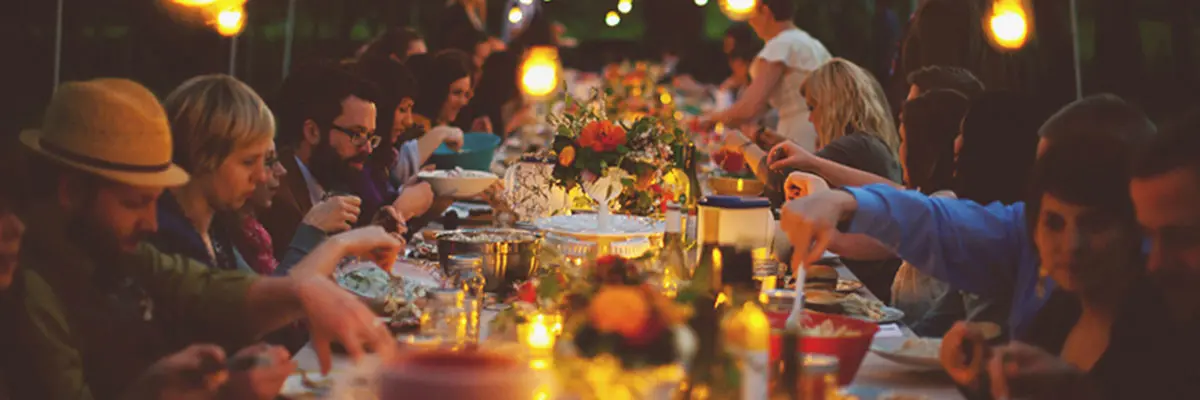  A long outdoor dinner table filled with people enjoying a meal, surrounded by decorative flowers and illuminated by warm string lights. 